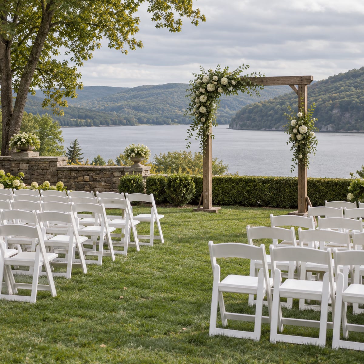 White chairs set in rows on an estate lawn facing a simple wooden arch, a river and wooded hills beyond