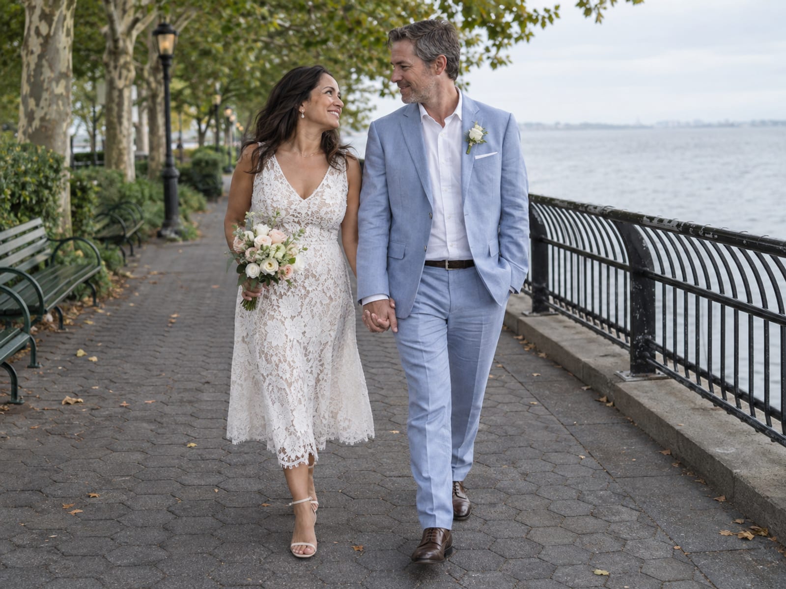 A woman with medium olive skin in a white sleeveless lace dress and a man with fair skin in a pale blue linen suit walk along a riverside park path of hexagonal pavers in soft early evening summer daylight, she carrying a small bouquet