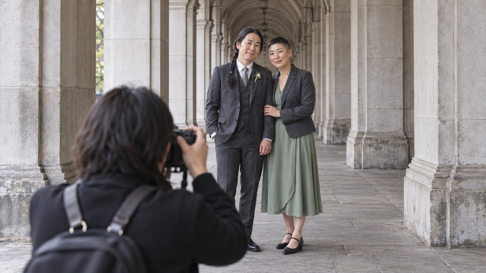 Two people in their forties, both with light skin, one in a charcoal three piece suit and one in a sage green dress with a tailored jacket, under a vaulted stone arcade between columns, a photographer in the foreground seen from behind with a camera raised
