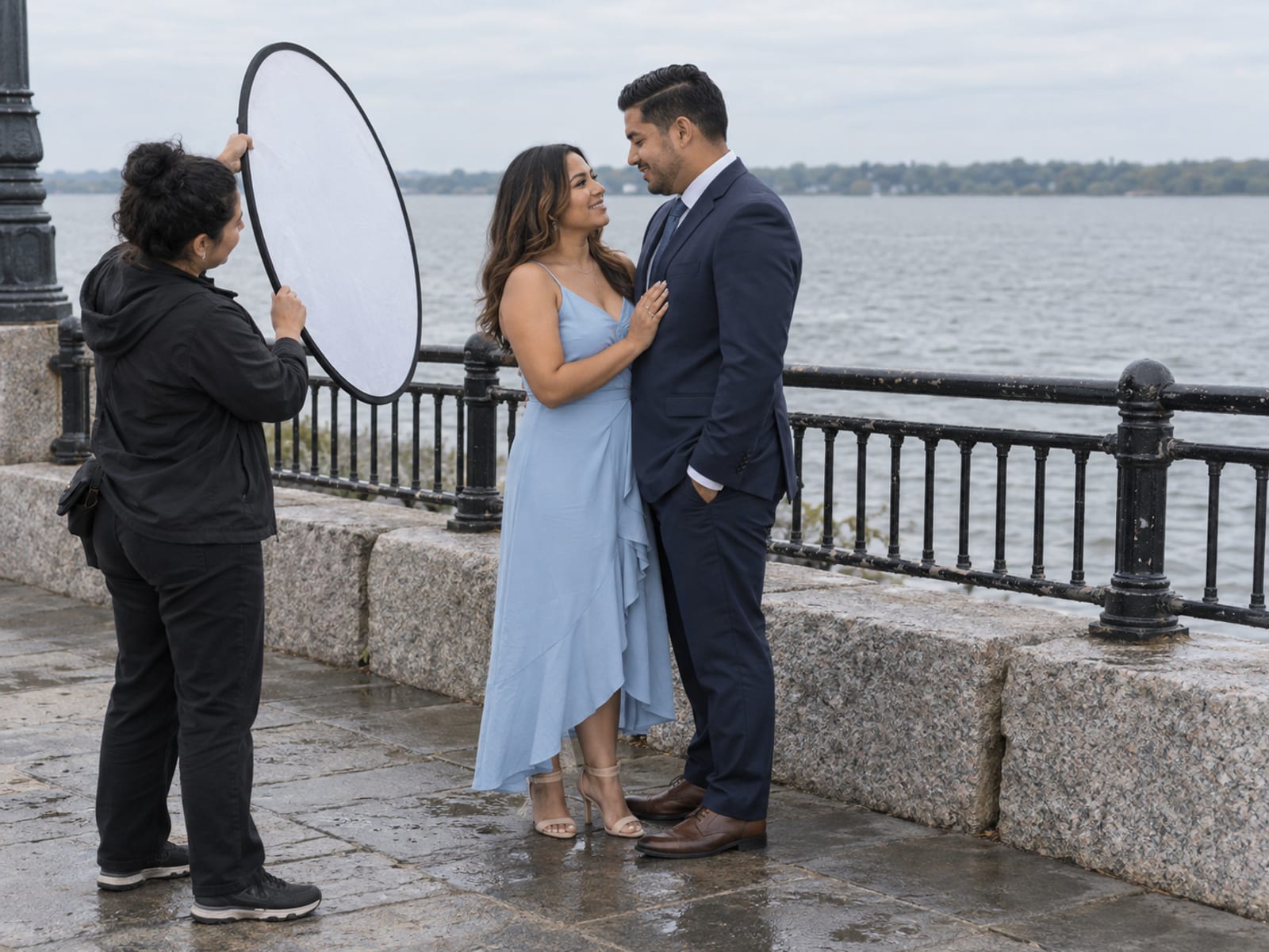 A photographer's assistant holding a round white reflector beside a woman in a sky blue dress and a man in a navy suit, both in their thirties with golden brown skin, on a waterfront esplanade of granite blocks and iron railings