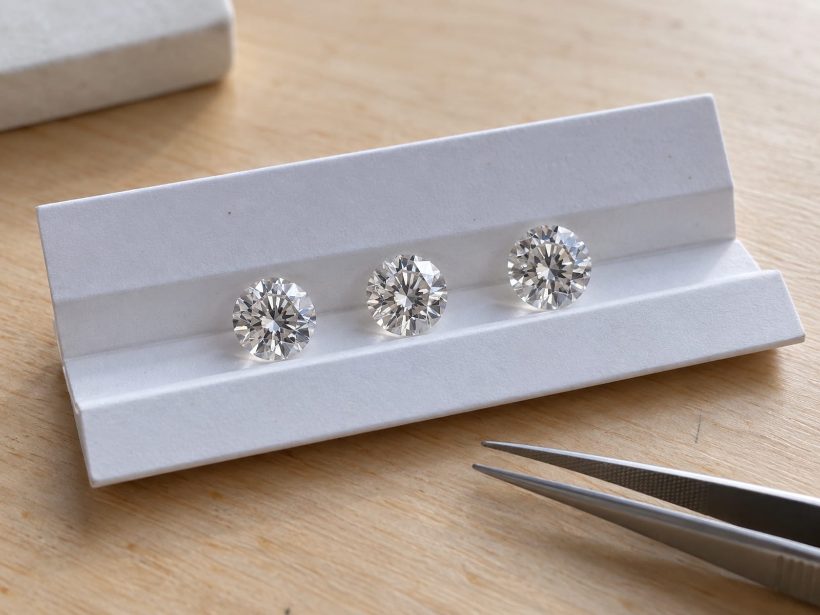 Three loose clear stones of similar size resting on a white grading tray beside fine steel tweezers on a light desk in soft north light
