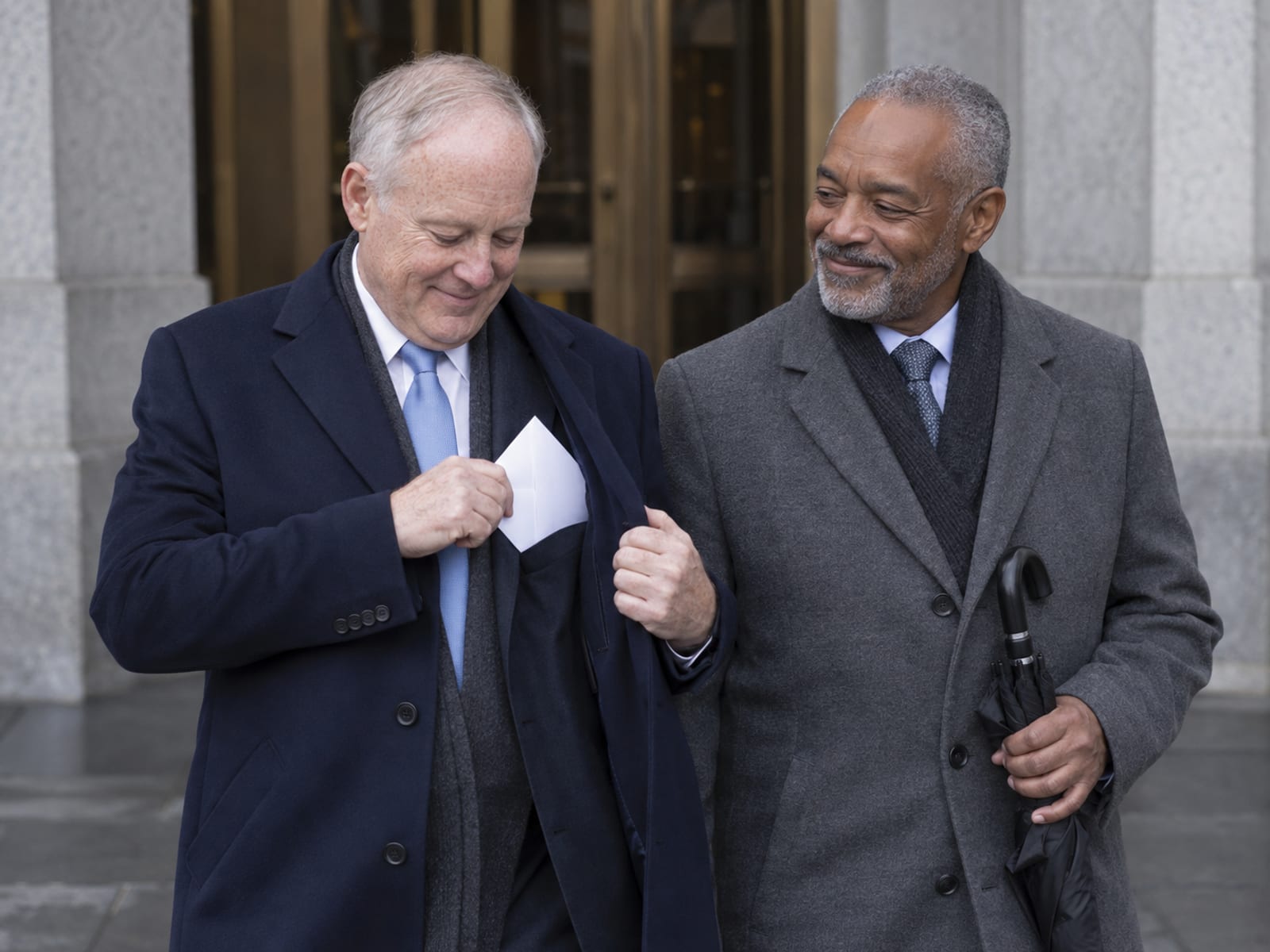 Two men in their sixties, one with fair freckled skin and one with medium brown skin, on a downtown sidewalk outside a granite civic building, one tucking a plain white envelope into an inside coat pocket while the other smiles holding a small umbrella