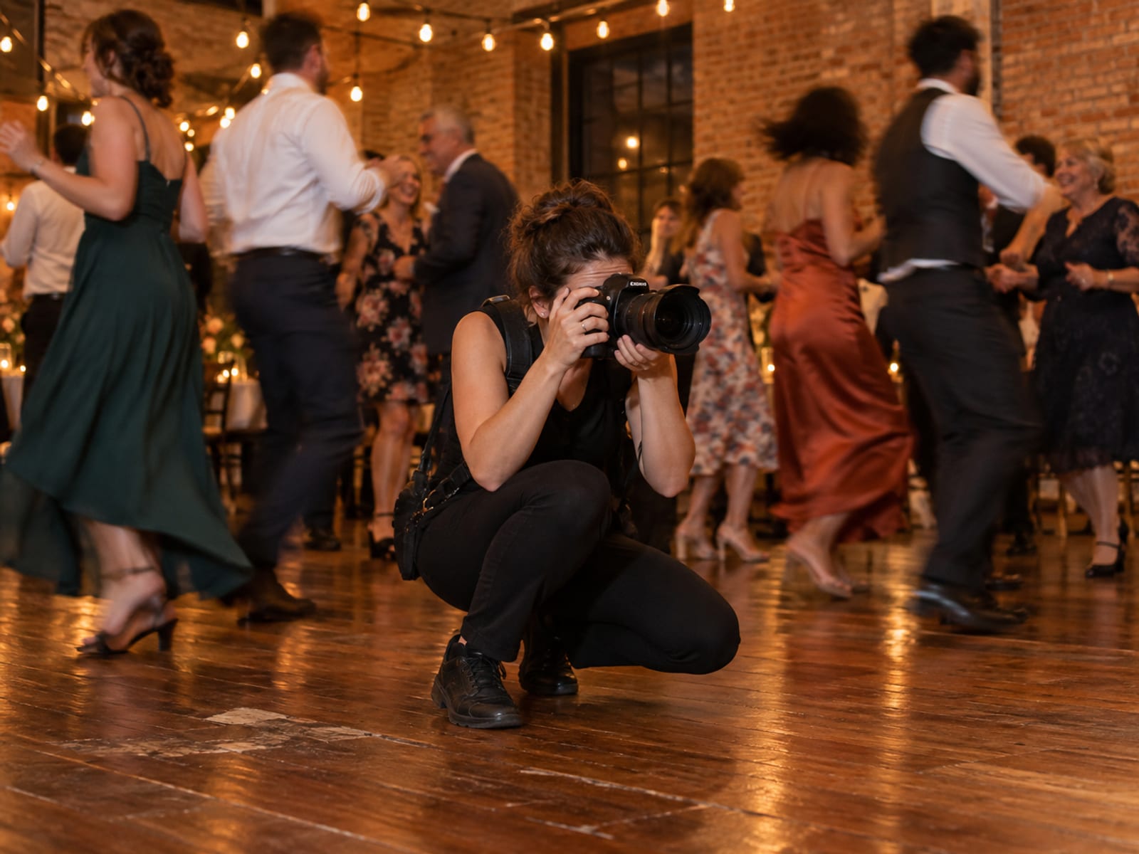 A photographer crouched low at the edge of a reception dance floor with the camera raised, guests of mixed ages dancing past in soft motion blur under strings of warm bulbs in a brick-walled room