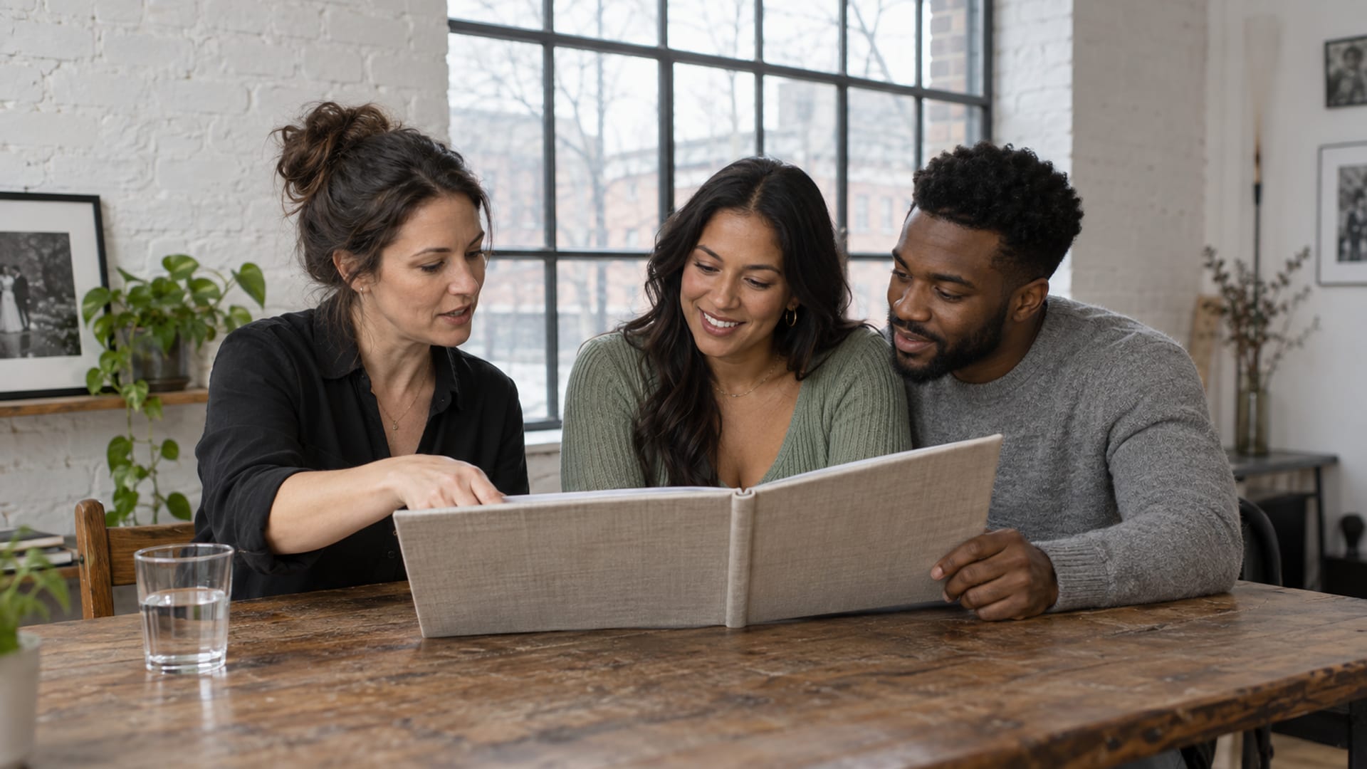 A photographer pointing to a page of a large linen bound album at a long wooden table in a whitewashed brick studio while a woman with warm tan skin and a man with dark brown skin lean in to turn the pages