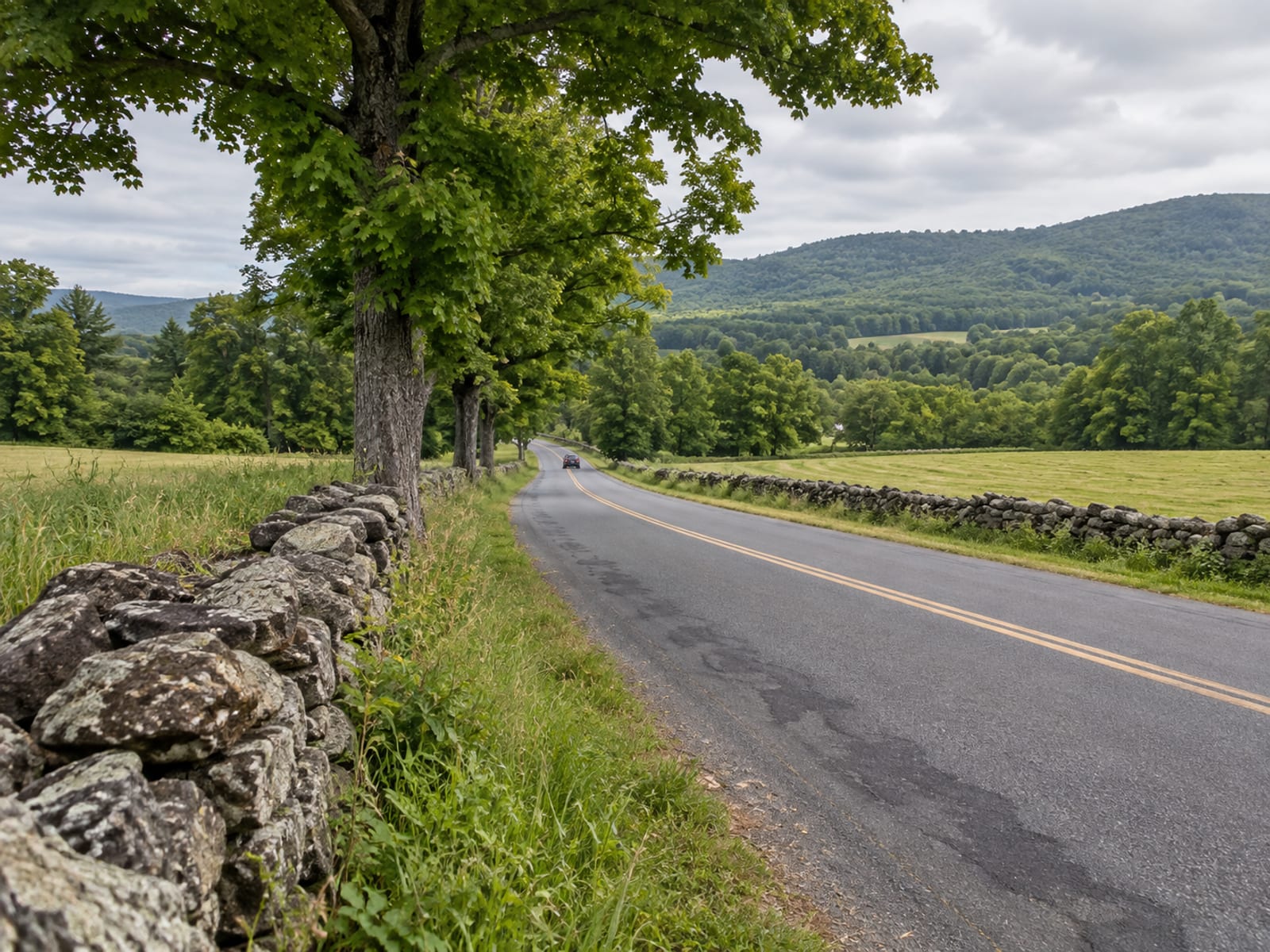 A narrow two-lane country road between fieldstone walls and hayfields leading toward wooded hills, one dark car small in the distance