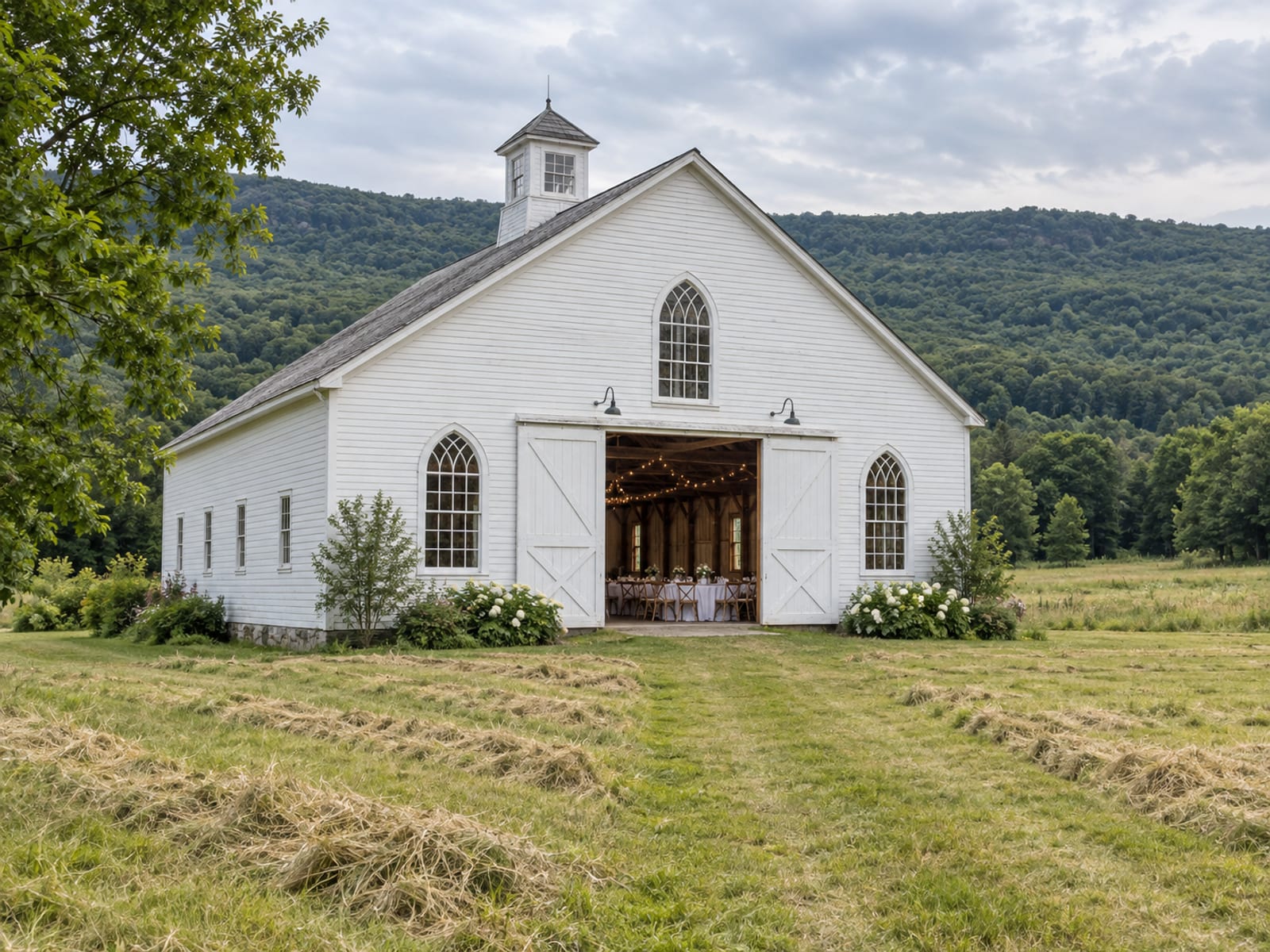 A large white clapboard barn with a cupola and arched windows in a mown hayfield, its doors open to show round tables set inside, wooded hills behind