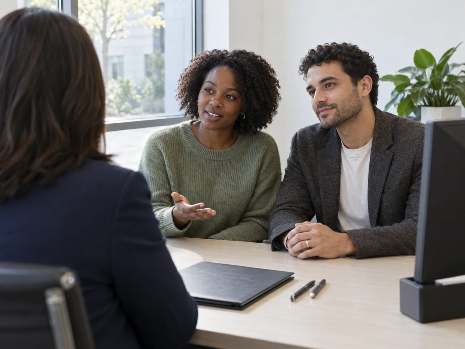 A woman and a man in their late twenties, one with deep brown skin and one with light olive skin, in everyday clothes seated side by side at a banker's desk in a plain bright bank branch, the banker seen from behind, a closed folder and two pens on the desk