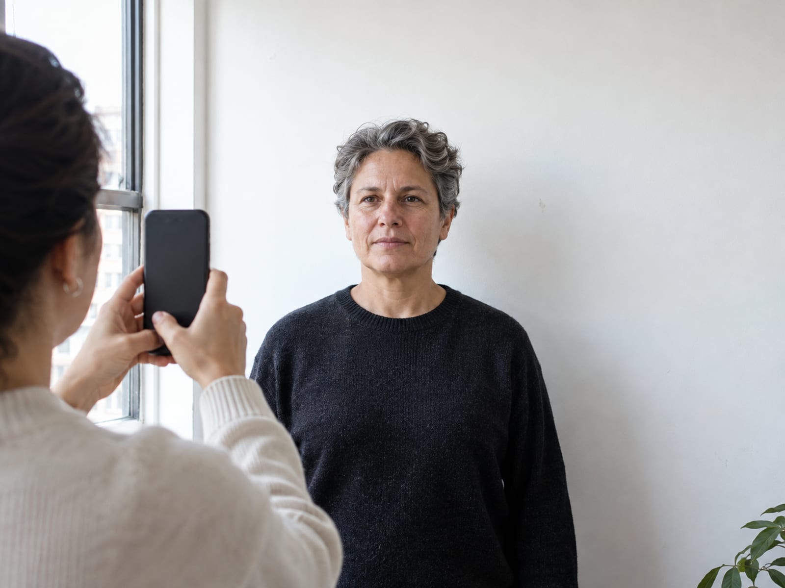 A person in their fifties standing against a plain white wall in an apartment while a friend takes their photo with a phone, the back of the phone toward the camera