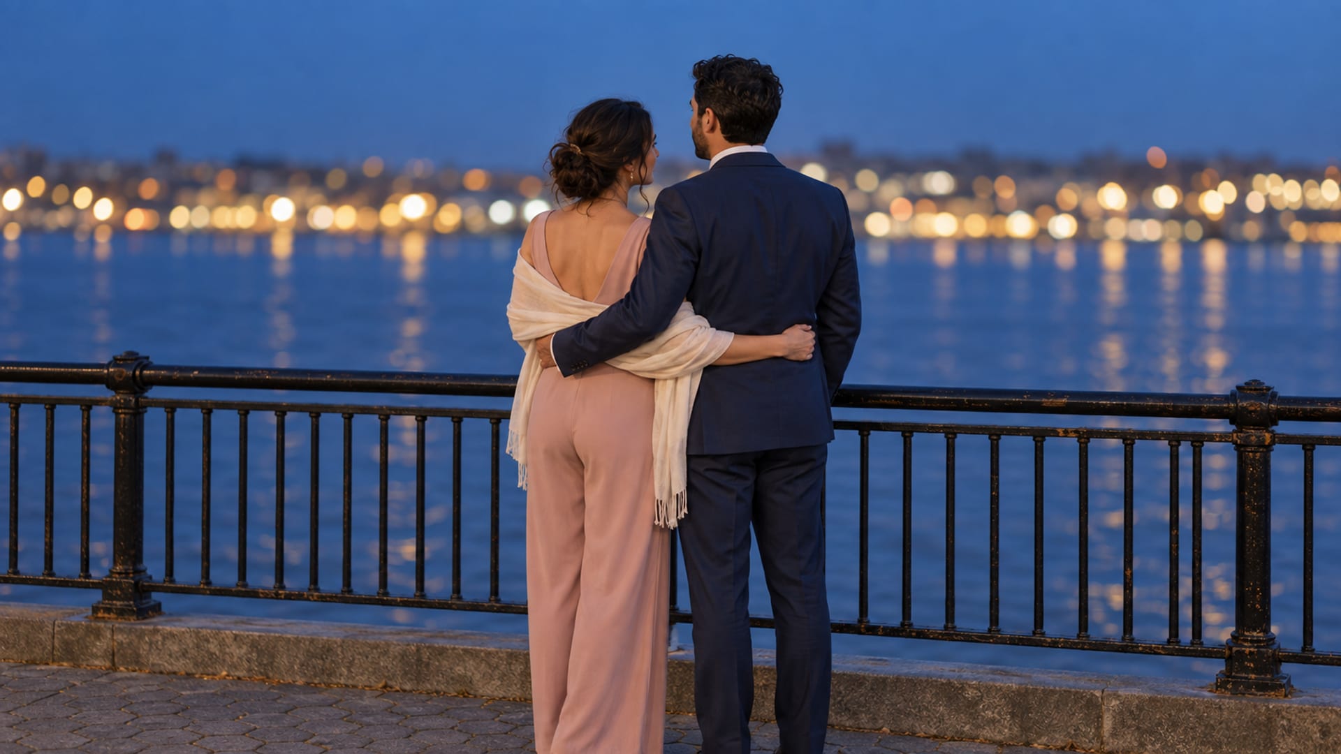 A woman in a blush jumpsuit and a man in a dark blue suit, both in their thirties with light olive skin, at the railing of a waterfront walkway at blue hour, the city across the river only a soft out-of-focus band of lights