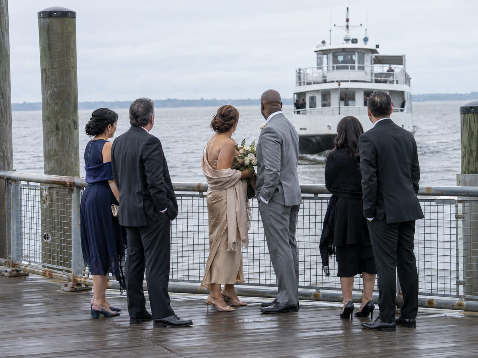 A small white commuter ferry pulling in to a wooden pier while a woman and a man in their fifties, one with warm tan skin and one with dark brown skin, wait at the railing with a few guests, one holding a bouquet