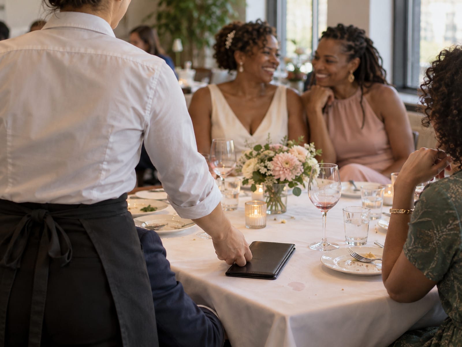 A server seen from behind setting a closed dark leather check folder on a white tablecloth at the end of a small wedding lunch, two women with dark brown skin and their guests out of focus