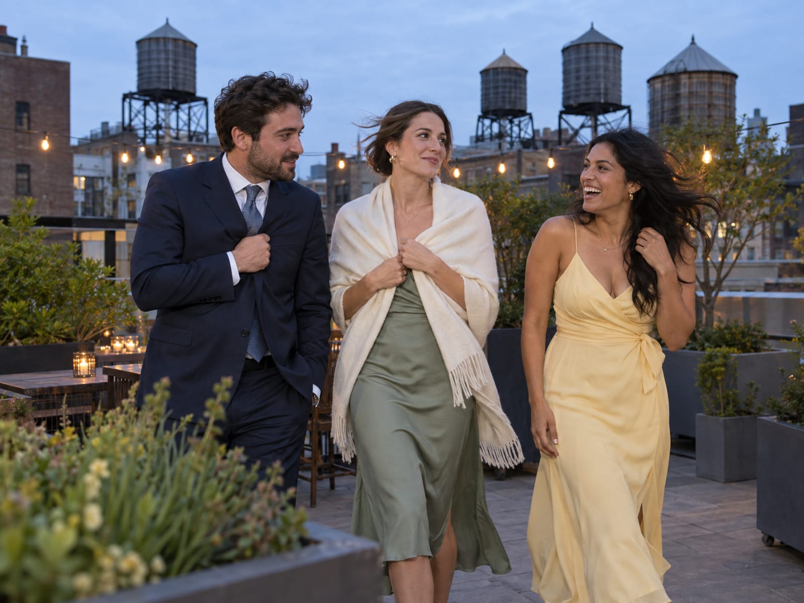 Wedding guests in their thirties on a windy rooftop terrace at dusk, one pulling a light wrap around the shoulders and another holding a jacket lapel against the wind, wooden water towers on neighboring roofs