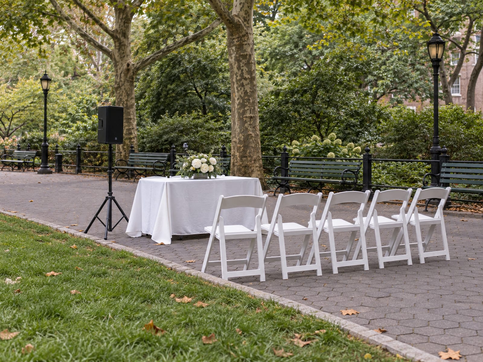 The edge of a park lawn beside a hexagonal-paver path readied for a ceremony: a folding table with a white cloth, a small speaker on a stand and a row of white chairs