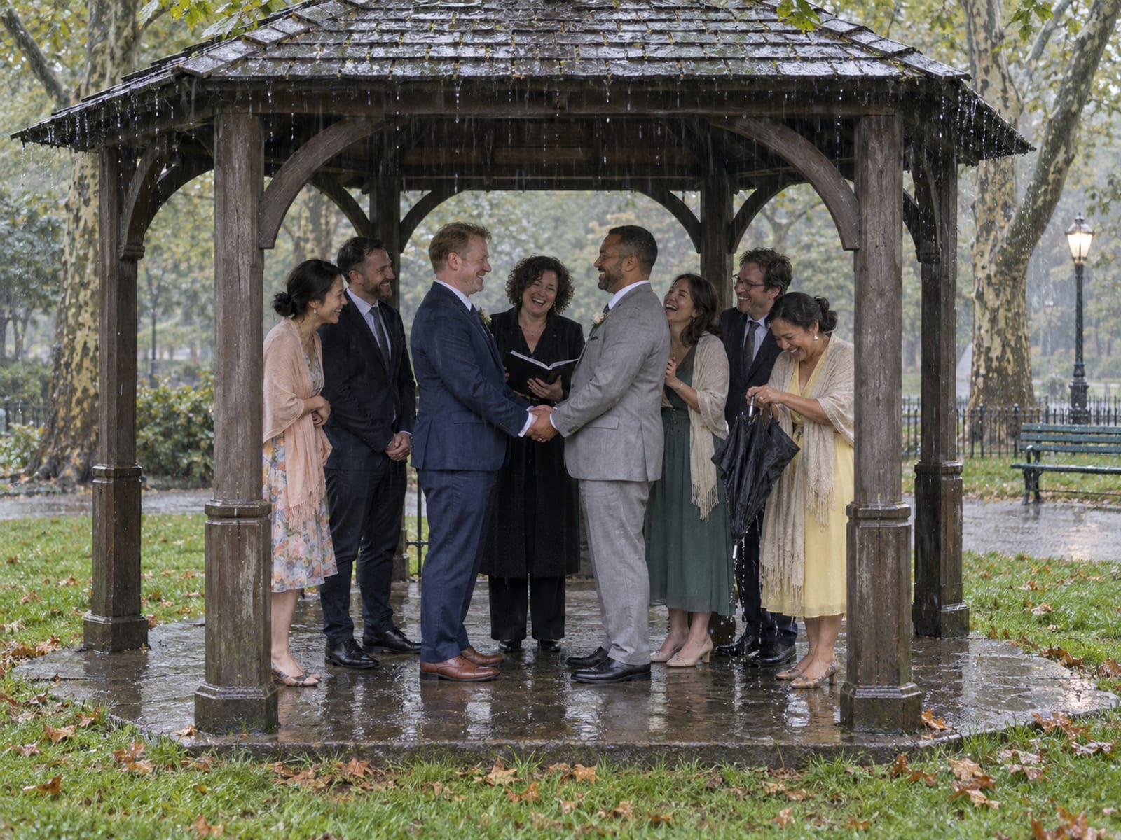 A ceremony group of eight sheltering under a small open wooden park pavilion in light rain, two men in their forties in navy and light grey suits, one with fair freckled skin and one with medium brown skin, and an officiant in the middle
