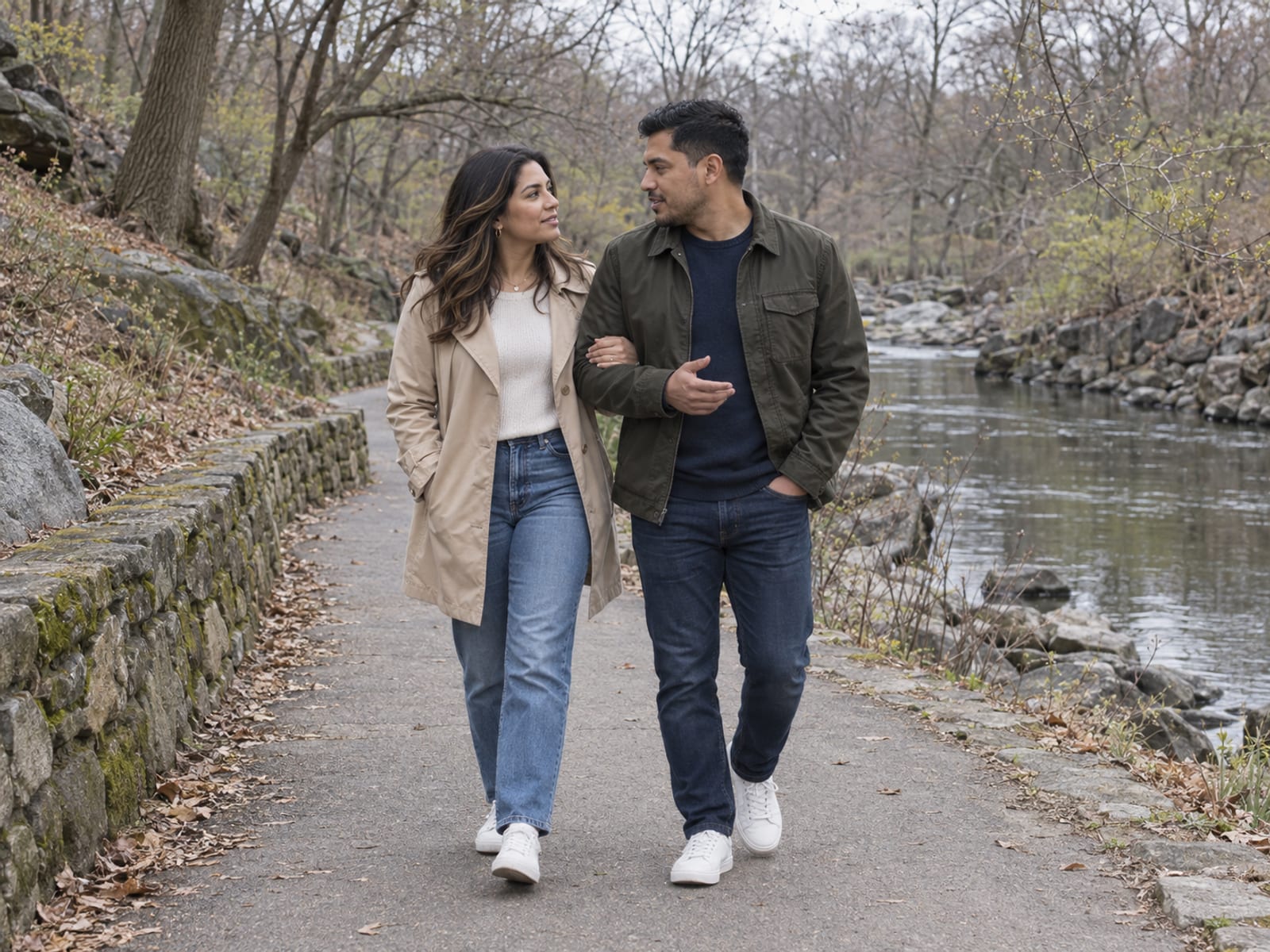 A woman and a man in their thirties, both with golden brown skin and Latin American features, walking slowly and talking on a quiet riverside path in a Bronx park, one with a hand on the other's arm, bare trees coming into bud