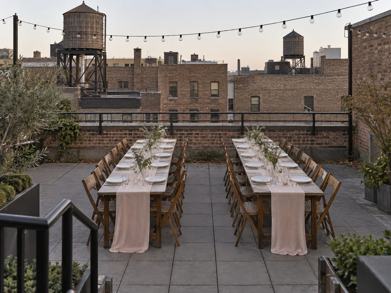 A Brooklyn rooftop terrace in early evening set with two long dinner tables under unlit string lights, a low parapet with a railing and wooden water tanks on neighboring roofs beyond