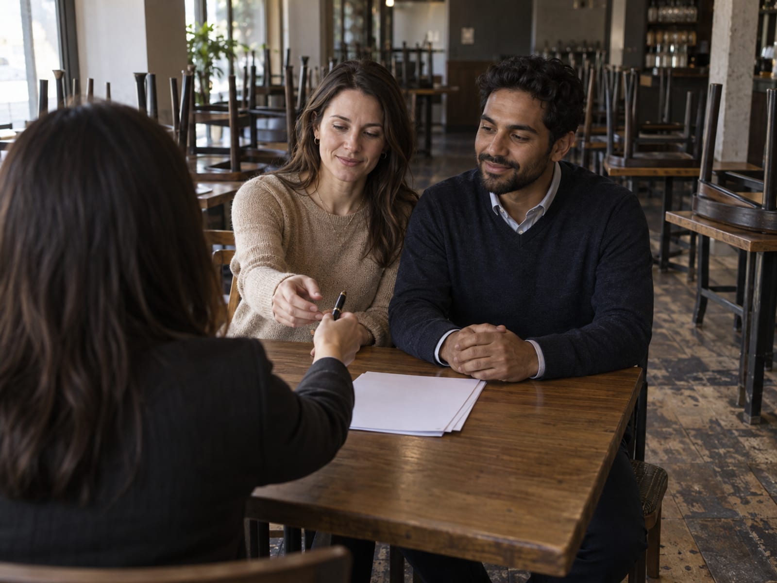 An events manager seen from behind handing a pen to a woman and a man in their thirties, one with pale skin and one with golden brown skin, as they sign a contract at a small table in an empty restaurant with chairs up on the tables behind