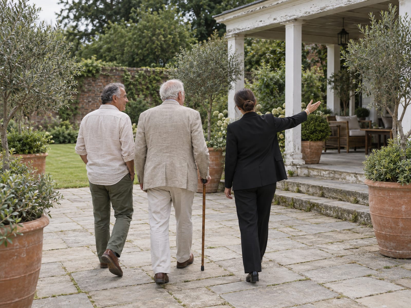 A venue coordinator seen from behind gesturing toward a covered wooden porch while walking two men in their sixties, one with medium olive skin and one with fair skin who walks with a wooden cane, across a stone terrace with potted olive trees