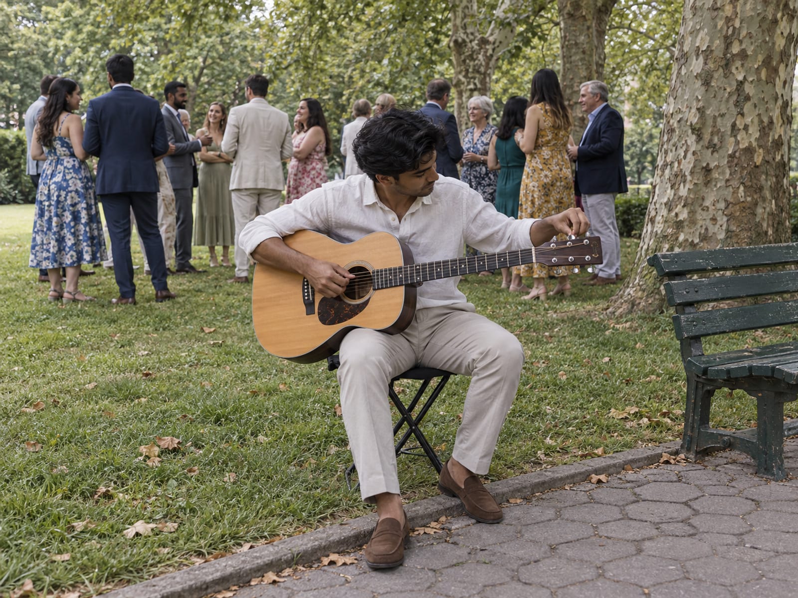 A friend in their twenties tuning an acoustic guitar on a folding stool at the edge of a Queens park lawn, about fifteen guests gathering on the grass nearby, no speakers or amplifier
