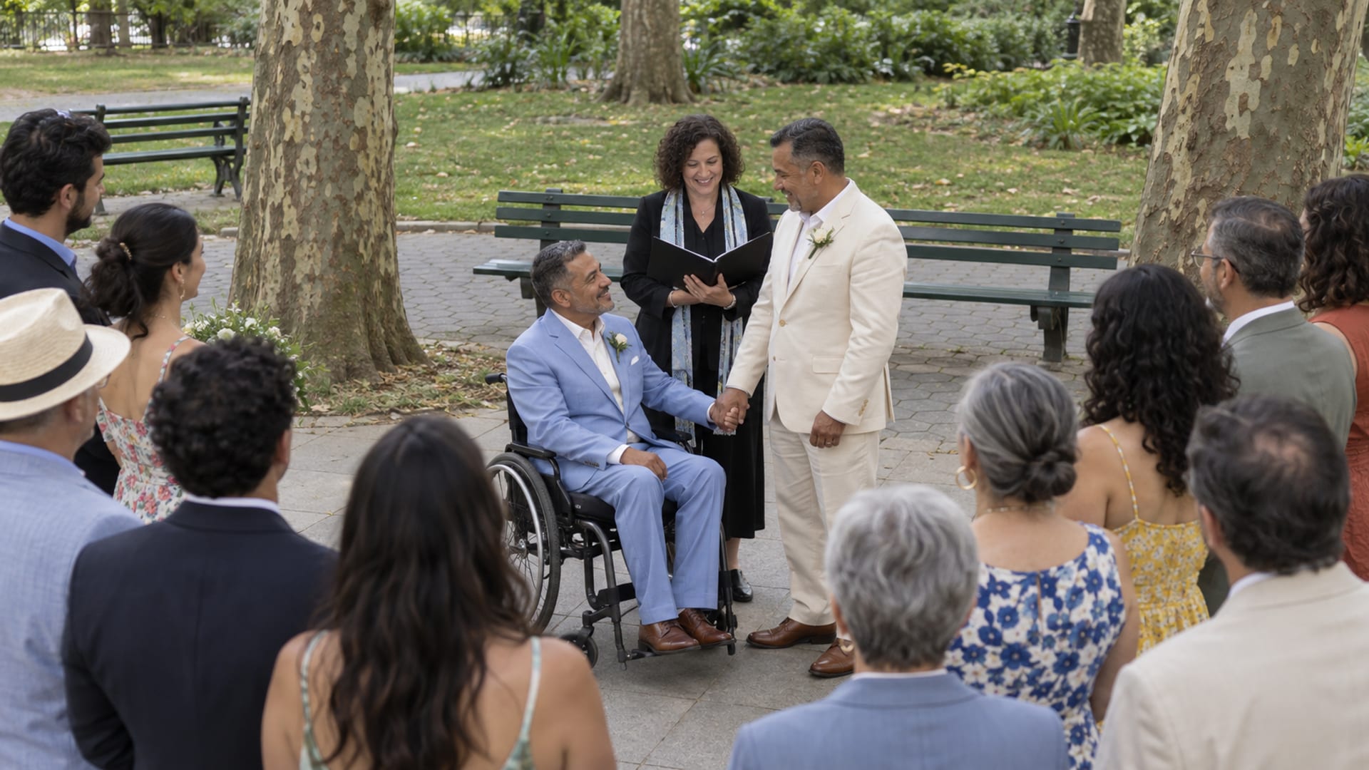 A small wedding ceremony at the edge of a park lawn under plane trees: two men in their fifties, both with golden brown skin, one in a light blue suit seated in a manual wheelchair and one in a cream suit, facing an officiant with about twelve guests in a loose semicircle