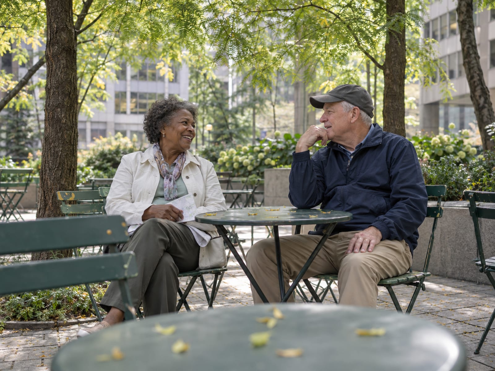 Two out-of-town guests in their sixties resting on movable metal chairs at a small table in a leafy Midtown public plaza under honey locust trees, one holding a folded paper map, office buildings softly blurred behind