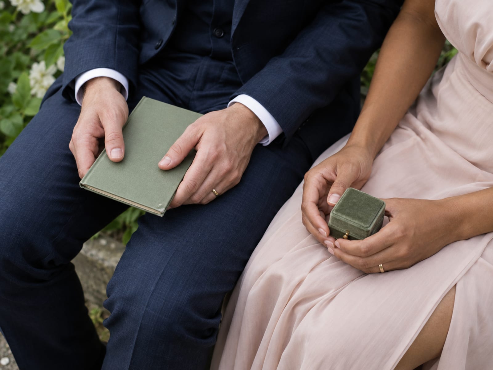 A close view of two people's hands in a garden, one holding a small vow card with a sturdy closed cover and the other holding a small ring box with a secure clasp, resting on a lap in formal clothes