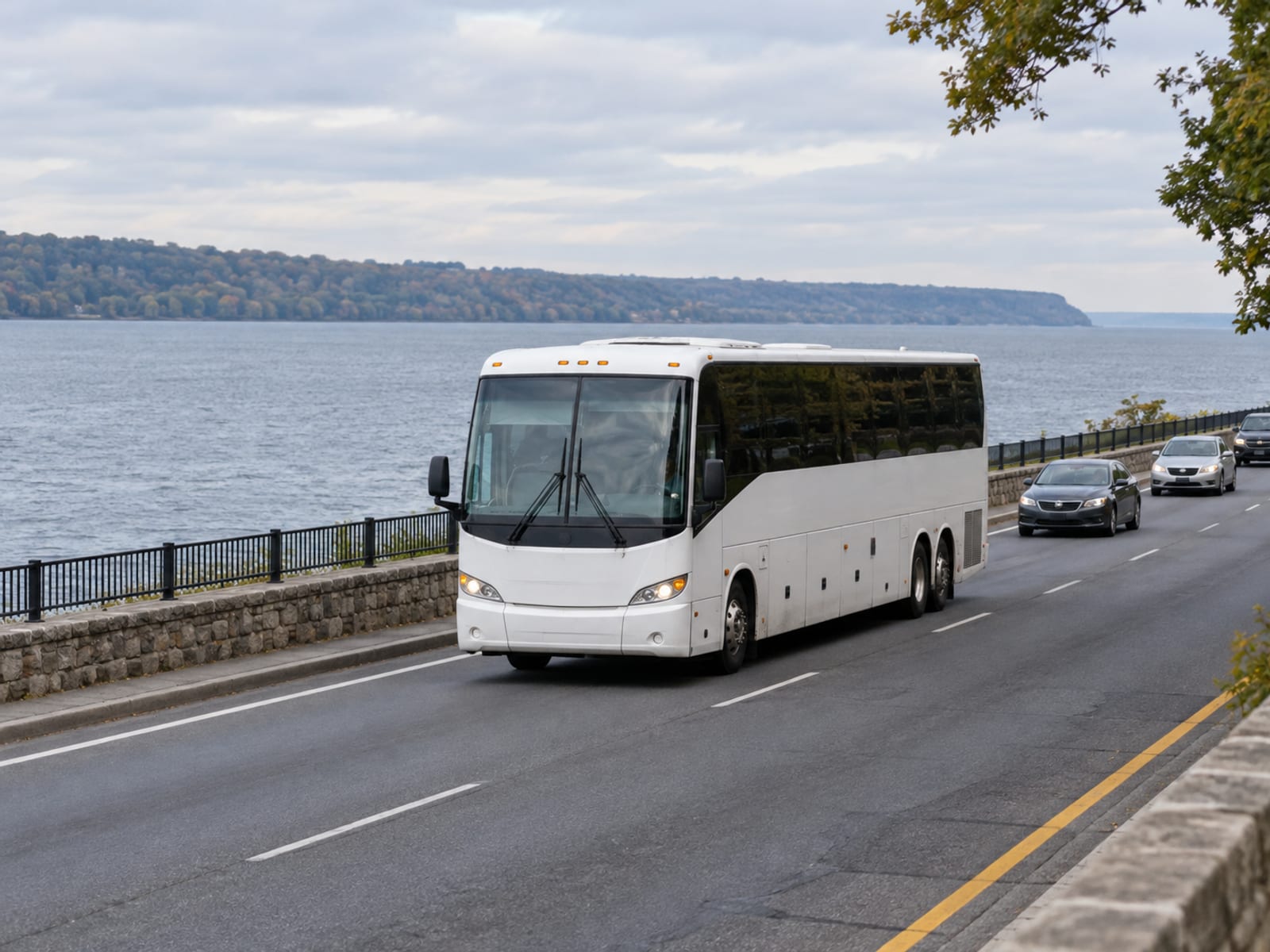 A white coach bus with no markings drives along a riverside highway in light morning traffic, with the river and a low far shore to one side