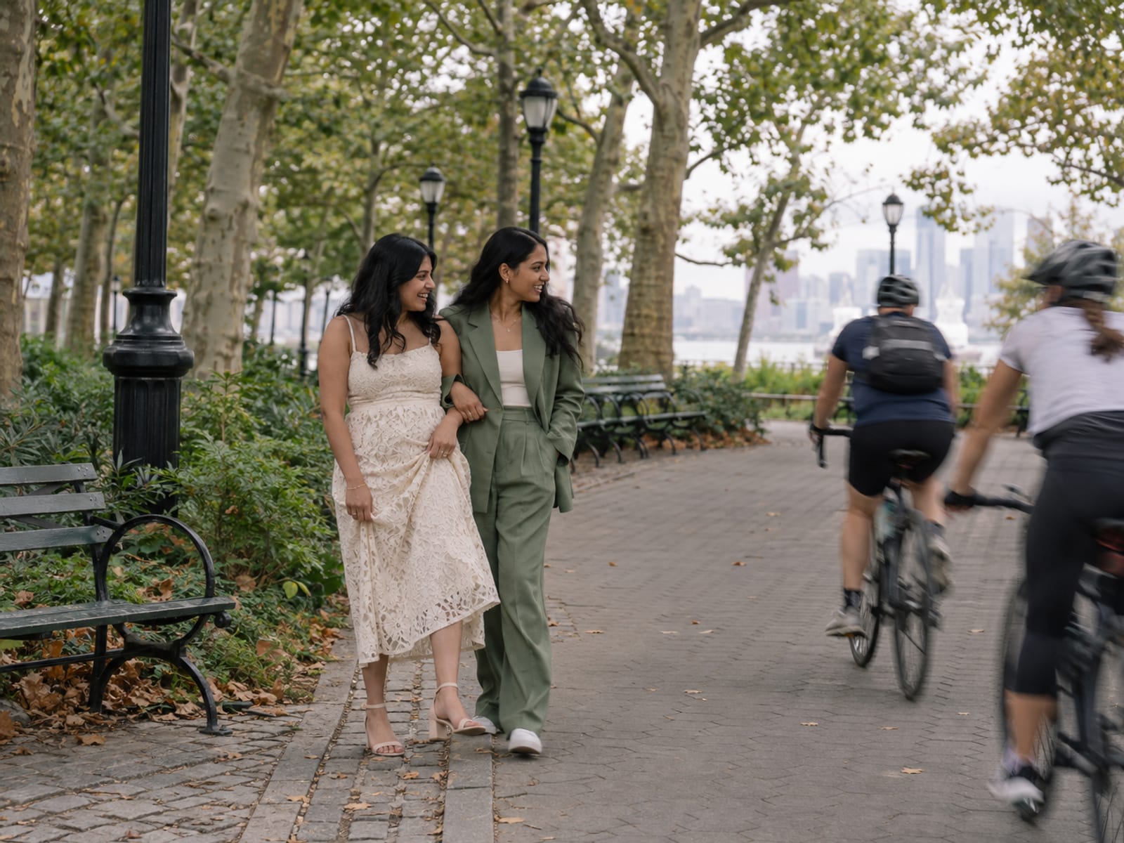 Two women in their twenties, both with warm tan skin, in a cream lace dress and a sage green suit, with a photographer, stepping to the edge of a wide park path while two cyclists ride past