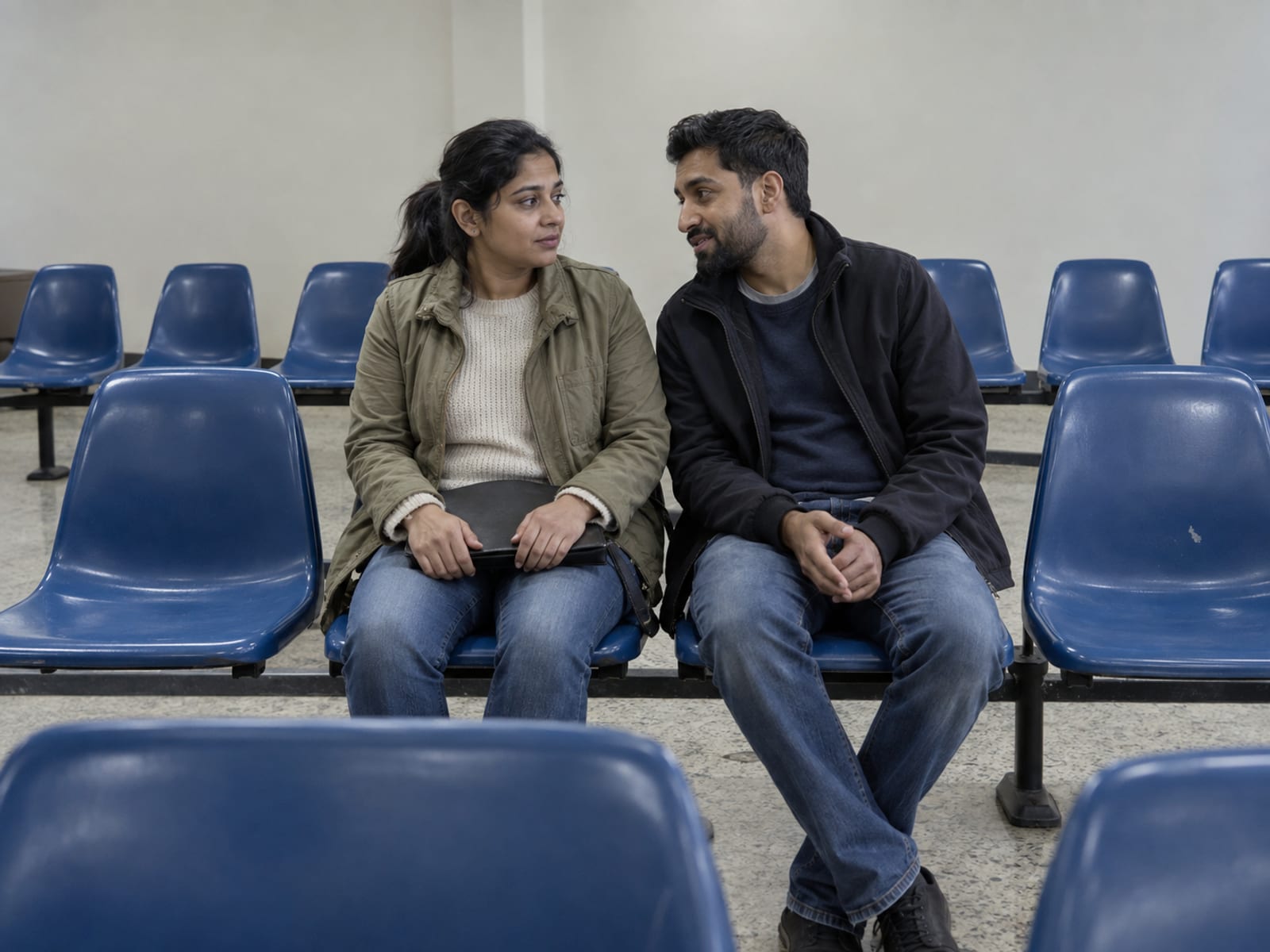 A woman and a man in their thirties, both with warm tan skin and South Asian features, seated side by side on plastic chairs in a plain government waiting room, one holding a closed dark folder on their lap, rows of empty chairs around them