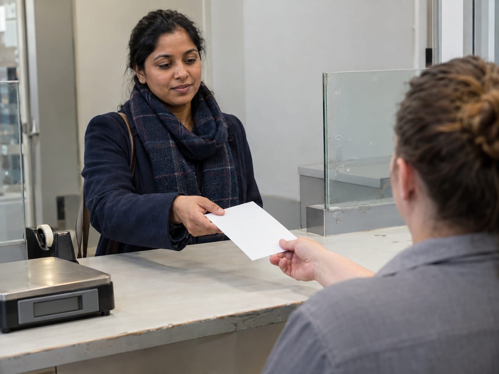 A person in their thirties at a plain post office counter handing a stiff white envelope to a clerk seen from behind, a parcel scale and a tape dispenser on the counter