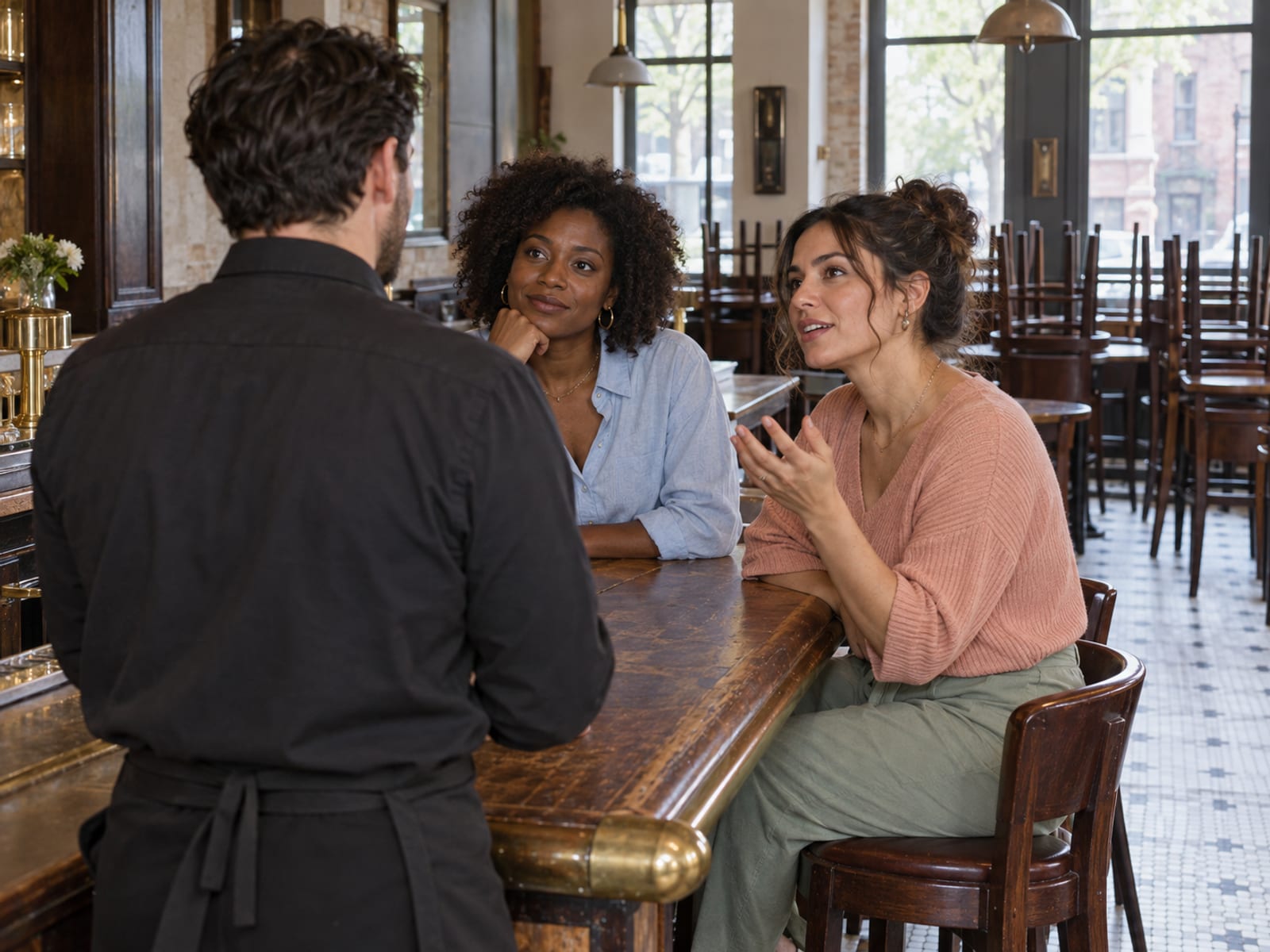 A restaurant events manager seen from behind talking with two women in their thirties, one with deep brown skin and one with light olive skin, at a bar counter on a quiet afternoon, chairs turned up on a few tables