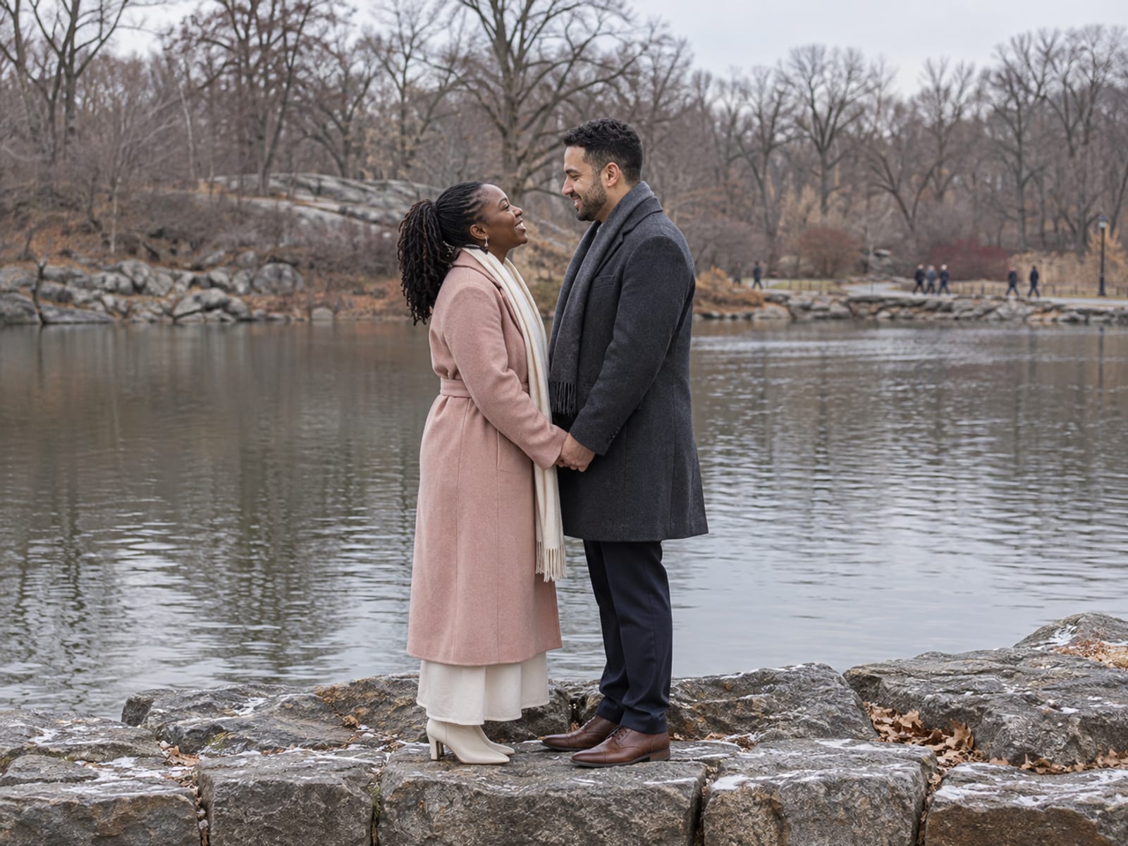 A woman and a man in their thirties, one with deep brown skin and one with light olive skin, hold hands and turn to face each other on a stone embankment beside a lake in a winter park