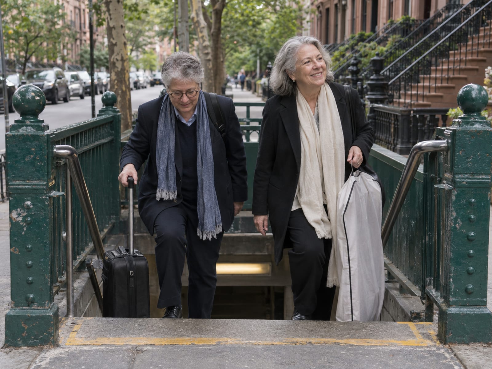 Dos invitados de boda de unos sesenta años con una maleta de ruedas pequeña suben las escaleras de una entrada del metro con barandas de hierro verde hacia una calle residencial arbolada