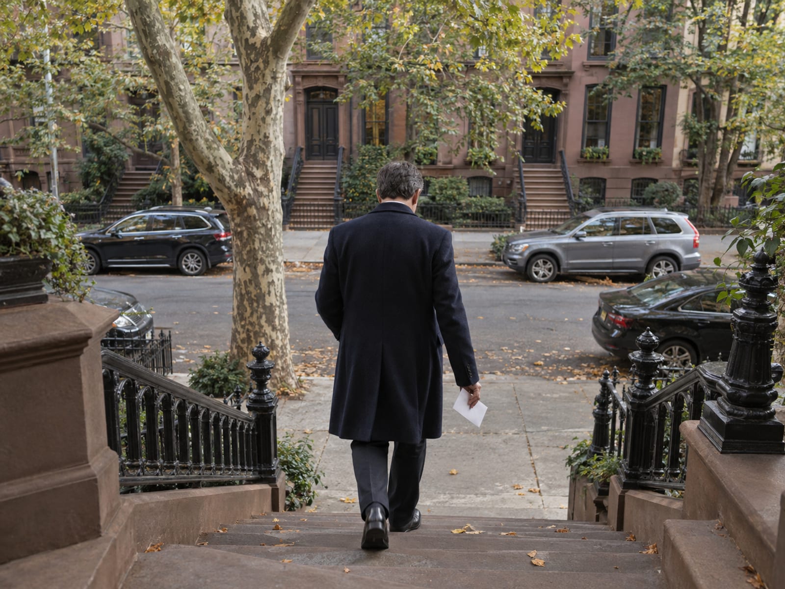 An officiant seen from behind walking down a brownstone stoop in the morning with a plain white envelope in one hand, the tree-lined block ahead