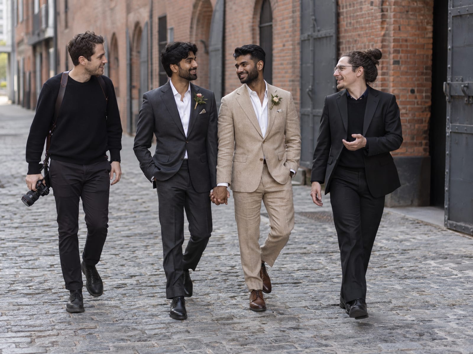 Two men in their twenties, both with warm tan skin, in charcoal and tan linen suits walking a cobbled lane between old brick buildings with an officiant and a photographer beside them