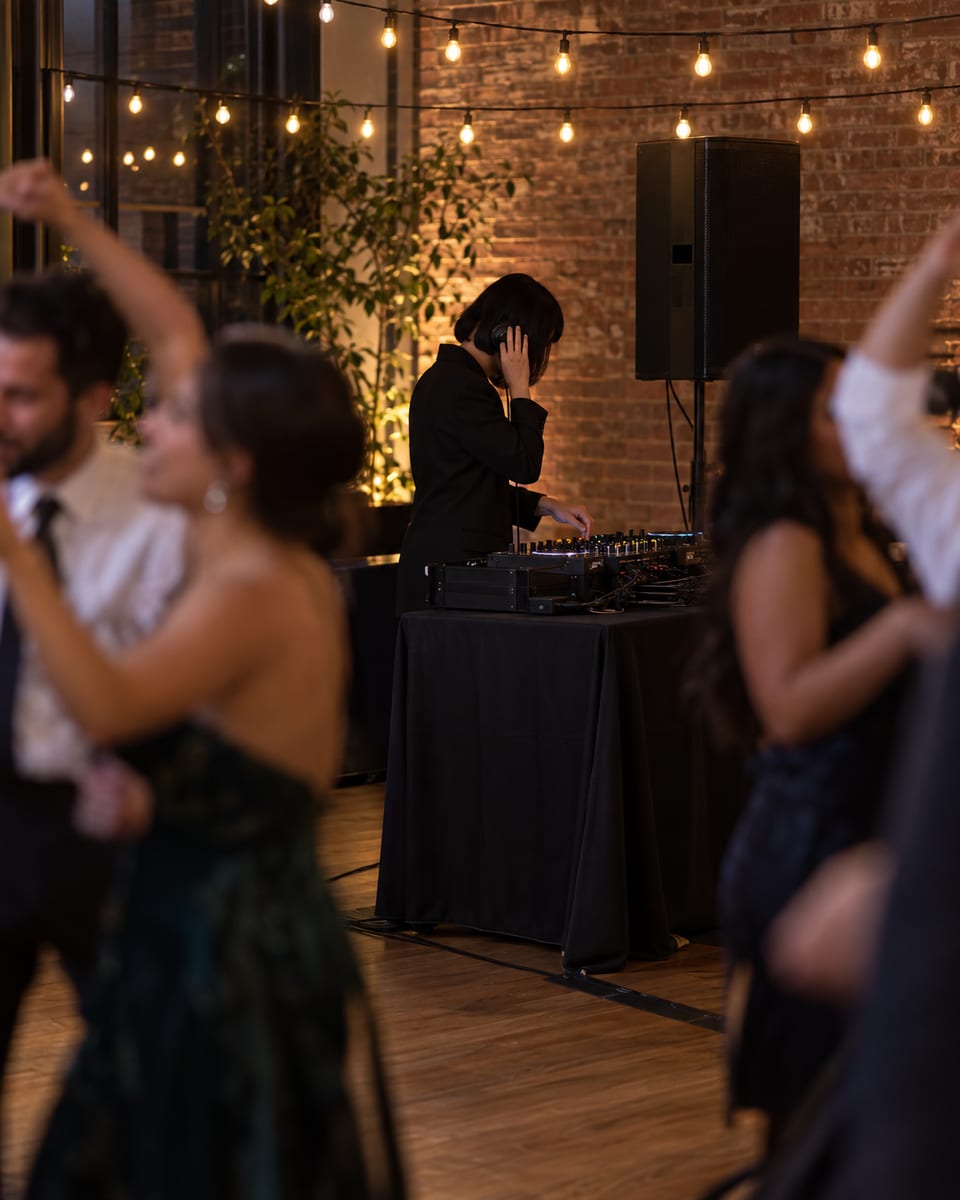 A DJ seen from the side, one headphone cup to an ear, cues music at a black-draped table beside a dance floor while guests dance blurred in the foreground